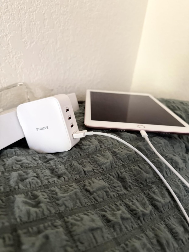 Philips 100W GaN charger plugged into wall socket neatly charging an iPad on bed